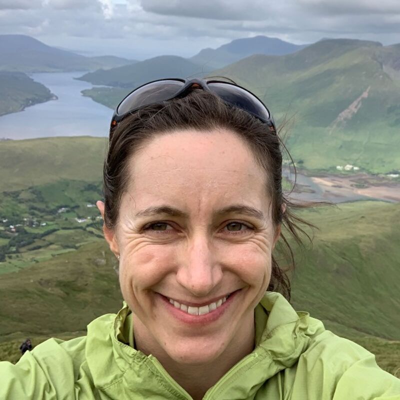 The image shows a woman smiling at the camera. She is wearing sunglasses and a green jacket. The background features a scenic view of mountains, a lake, and a cloudy sky. The woman appears to be enjoying the outdoors, possibly hiking or exploring a mountainous region.